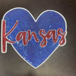 May include: A heart-shaped patch with a blue sequined background and the word "Kansas" in red sequins. The word is outlined in white. The patch is likely for apparel or accessories.