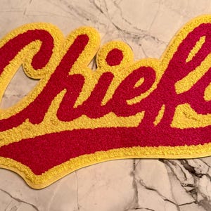 May include: A yellow and red embroidered patch with the word "Chiefs" in a script font. The word is in red with a yellow outline. The patch is on a marble surface.