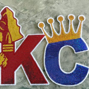 May include: Red and blue felt letters "KC" with a gold crown on top of the "C". The "K" has a gold spearhead design on the left side.