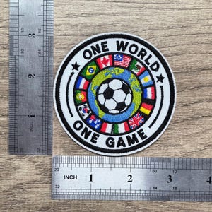 May include: A circular embroidered patch featuring a central soccer ball over a globe, surrounded by various national flags and the text "ONE WORLD ONE GAME". A metal ruler shows the patch has a diameter of approximately 3 inches.