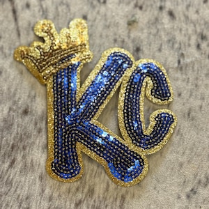 May include: A gold glitter crown sits atop the letters "KC" in a patch. The letters are filled with blue sequins and outlined in gold glitter. The patch is on a textured, neutral-toned surface.