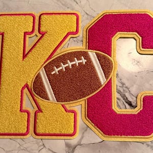 May include: A yellow letter "K" and a pink letter "C" with a brown football in the center. The letters and football have a textured appearance with a red outline and a light tan border.