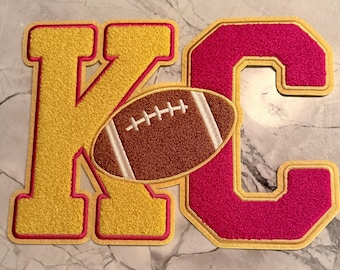 May include: A yellow letter "K" and a pink letter "C" with a brown football in the center. The letters and football have a textured appearance with a red outline and a light tan border.