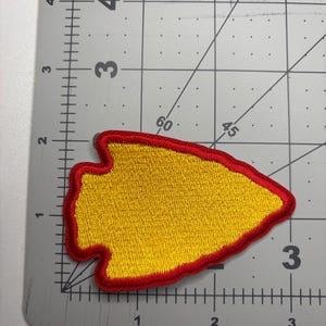 May include: Embroidered patch in the shape of a yellow arrowhead with a red border. The patch is approximately 3 inches long and 2 inches wide. The patch is on a gray cutting mat with measurement markings.