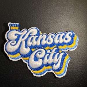 May include: A white embroidered patch with the words "Kansas City" in a retro, wavy font. The letters are outlined in blue and yellow, and a small crown sits above the word "Kansas".