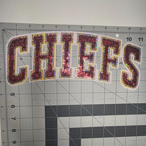 May include: A sequined patch with the word "CHIEFS" in maroon and gold lettering. The letters are outlined in gold and filled with shimmering sequins. The patch is on a white background.