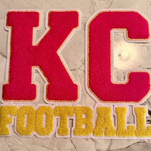 May include: Red and gold felt patches spelling out "KC FOOTBALL". The letters "K" and "C" are in a bold, blocky font with a white border and gold trim. The word "FOOTBALL" is in gold with a white border.