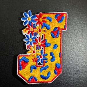 May include: Embroidered patch in the shape of the letter "J". The patch features a yellow background with blue and red leopard print spots. A cluster of embroidered flowers in blue, red, and yellow adorns the left side.