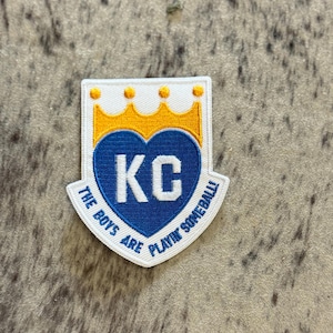 May include: White embroidered patch with a blue heart containing the letters "KC" and a yellow crown above the heart. The text "The Boys Are Playin' Someball" is written around the heart.