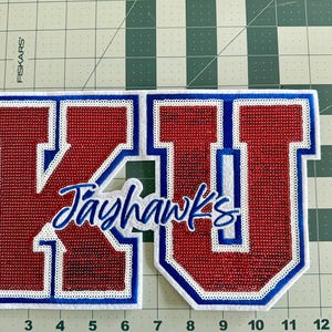 May include: Red and blue felt patch with the letters "KU" in white outlined in blue. The word "Jayhawks" is written in blue cursive below the letters.