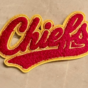 May include: A red and gold embroidered patch with the word "Chiefs" in a script font. The letters are a vibrant red with a gold outline, creating a bold and eye-catching design. The patch has a textured, fuzzy appearance.
