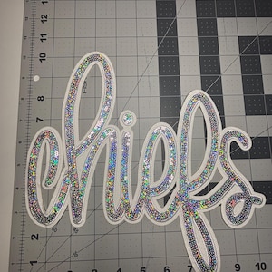 May include: Two white, sequined "Chiefs" script patches. The letters are outlined in silver and filled with iridescent sequins. The patches are on a gray cutting mat with grid lines.