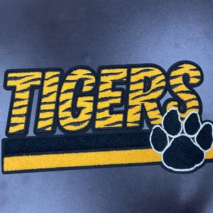 May include: A black, yellow, and white patch with the word "TIGERS" in yellow tiger stripe lettering. A black paw print with white outlines is on the right. A black and yellow stripe is below the word.