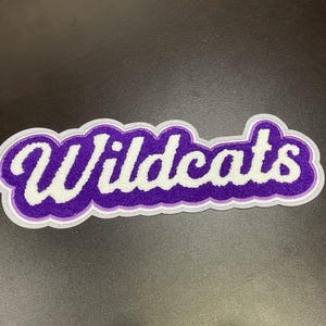 May include: A purple and white Wildcats patch with a white border. The word "Wildcats" is in a white, cursive font with a purple outline. The patch has a felt-like texture and is on a dark surface.
