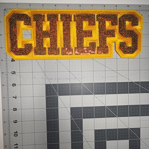 May include: A yellow and red sequined patch with the word "CHIEFS" in large, block letters. The letters are outlined in yellow and filled with red sequins. The patch is rectangular with rounded corners.