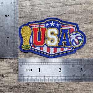 May include: An embroidered patch featuring the text USA in red, white, and blue, flanked by a gold trophy and a soccer ball. The patch is positioned against two metal rulers, showing a width of approximately 3.5 inches and a height of 2 inches.