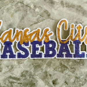 May include: Kansas City Baseball patch with gold and blue sequins. The patch is white with a white border.
