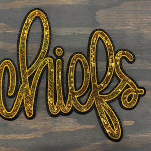 May include: A gold sequined patch with the word "Chiefs" in a cursive font. The patch is outlined in black.