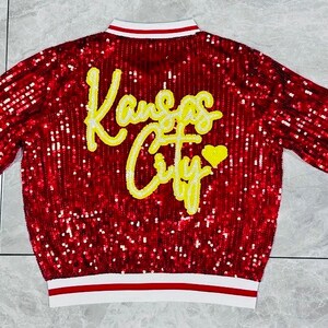 K&ouml;nnte beinhalten: Rote Pailletten-Bomberjacke mit wei&szlig;-rot gestreiften B&uuml;ndchen und Kragen. Auf dem R&uuml;cken steht in gelber Schrift "Kansas City" mit einem gelben Herz. Die Jacke ist ein auff&auml;lliges, auff&auml;lliges Kleidungsst&uuml;ck.