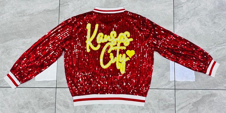 K&ouml;nnte beinhalten: Rote Pailletten-Bomberjacke mit wei&szlig;-rot gestreiften B&uuml;ndchen und Kragen. Auf dem R&uuml;cken steht in gelber Schrift "Kansas City" mit einem gelben Herz. Die Jacke ist ein auff&auml;lliges, auff&auml;lliges Kleidungsst&uuml;ck.