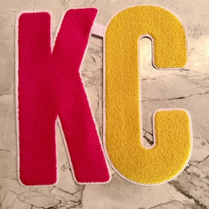 May include: A pair of varsity-style patches spelling "KC". The letter "K" is a vibrant pink, and the "C" is a bright yellow. Both letters have a fuzzy texture and are outlined with a white border. The patches are likely for apparel or crafts.