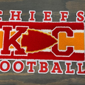May include: A red, white, and gold patch with the words "CHIEFS FOOTBALL". The patch features a red arrowhead and the letters "KC" in red and gold. The patch is likely for a sports team.