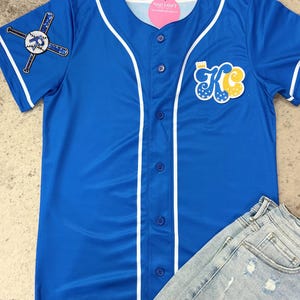 Op de afbeelding: Een koninklijk blauw baseballshirt met witte afwerking en een witte knoopsluiting. Het shirt heeft een groot "KC"-logo op de voorkant met sterren en een kroon. De linkermouw heeft een afbeelding van een honkbalknuppel en een bal.