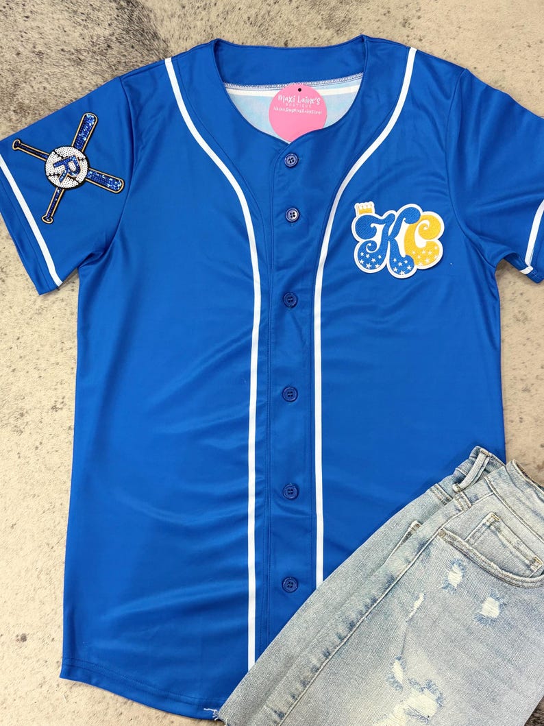 Op de afbeelding: Een koninklijk blauw baseballshirt met witte afwerking en een witte knoopsluiting. Het shirt heeft een groot "KC"-logo op de voorkant met sterren en een kroon. De linkermouw heeft een afbeelding van een honkbalknuppel en een bal.