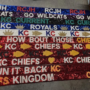 May include: A collection of colorful beaded banners with various text, including "RCJH", "GO WILDCATS", "KC CURRENT", "KC ROYALS", "HOW BOUT THOSE CHIEFS", "KC CHIEFS", "RUN IT BACK", and "CHIEFS KINGDOM".