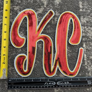 May include: A red sequined patch in the shape of the letters KC with a white and yellow border. A yellow measuring tape shows a height of approximately 8 inches, and a black ruler shows a width of approximately 8 inches.