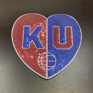 May include: Heart-shaped decorative item, split into red and blue sections. The letters "KU" are prominently displayed, with a basketball graphic below. The design is made of small, sparkling beads.