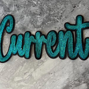 May include: A turquoise blue sequin patch with the word "Current" in a cursive font. The patch is outlined in black.