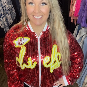 K&ouml;nnte beinhalten: Rote Pailletten-Bomberjacke mit wei&szlig;er und gelber Schrift &bdquo;Chiefs&ldquo;. Die Jacke hat einen wei&szlig;en Rei&szlig;verschluss und rot-wei&szlig; gestreifte B&uuml;ndchen und Kragen. Die Jacke ist ein auff&auml;lliges Kleidungsst&uuml;ck.