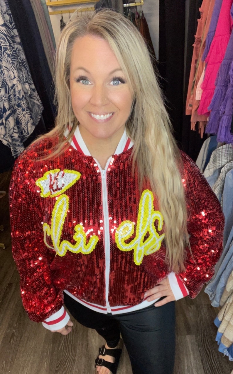 K&ouml;nnte beinhalten: Rote Pailletten-Bomberjacke mit wei&szlig;er und gelber Schrift &bdquo;Chiefs&ldquo;. Die Jacke hat einen wei&szlig;en Rei&szlig;verschluss und rot-wei&szlig; gestreifte B&uuml;ndchen und Kragen. Die Jacke ist ein auff&auml;lliges Kleidungsst&uuml;ck.