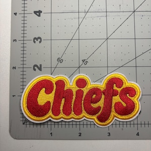 May include: Embroidered patch featuring the word "Chiefs" in red, outlined in yellow and white. The patch is rectangular and has a stitched design. The patch is on a gray cutting mat.