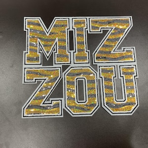 May include: A set of sequined patches spelling out "MIZ ZOU" in gold, black, and white. The letters have a tiger stripe pattern and are outlined in black and white. The patches are likely for apparel.