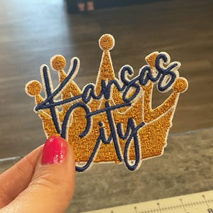 May include: Embroidered patch in the shape of a gold crown with the words "Kansas City" in navy blue script. The patch has a textured appearance and is likely designed to be ironed or sewn onto fabric.