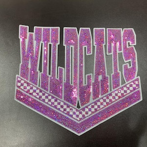 May include: A purple and pink rhinestone patch with the word "WILDCATS" in a bold, stylized font. The patch has a white border and a checkered pattern at the bottom. The rhinestones sparkle, creating a glamorous effect.