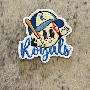 May include: White embroidered patch with a cartoon baseball character wearing a blue hat with the letter "R" on it. The character is holding a wooden bat and has a smiling face. The text "Royals" is embroidered in blue below the character.