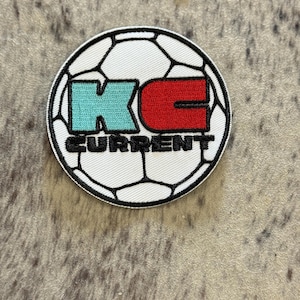 May include: A white soccer ball patch with black stitching. The letters "KC" are in the center of the patch, with "K" in turquoise and "C" in red. The word "CURRENT" is written in black below the letters.