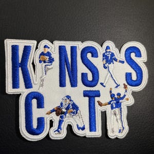 May include: A white embroidered patch with the words "KANSAS CITY" in blue lettering. The patch features five baseball players in blue uniforms.