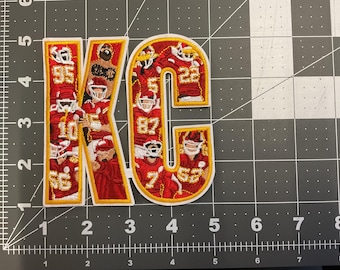 May include: Embroidered patch featuring the letters "KC" in red and gold, filled with images of football players in uniform. The patch has a white border and is placed on a gray grid.