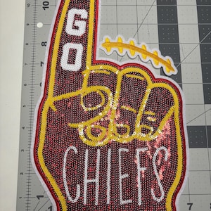 May include: A red, yellow, and white foam finger patch with the words "GO CHIEFS" in white and gold sequins. The patch has a yellow outline and a white border. A yellow and white football is attached.