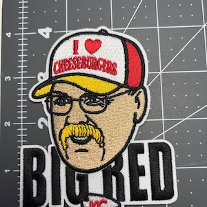 May include: Embroidered patch featuring a cartoon image of a man wearing glasses and a baseball cap. The cap reads "I ❤️ CHEESEBURGERS." Below the image is the text "BIG RED" and a "KC" logo.