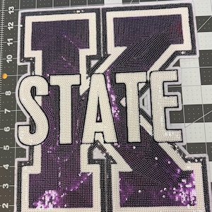 May include: Purple and white sequined iron-on patch with the word "STATE" and a large letter "K".