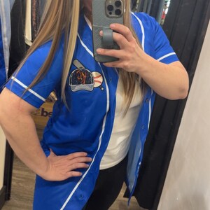 Op de afbeelding: Een blauw baseballshirt met witte bies en een baseball-thema patch op de voorkant. Het shirt is knoopsluiting en heeft korte mouwen.