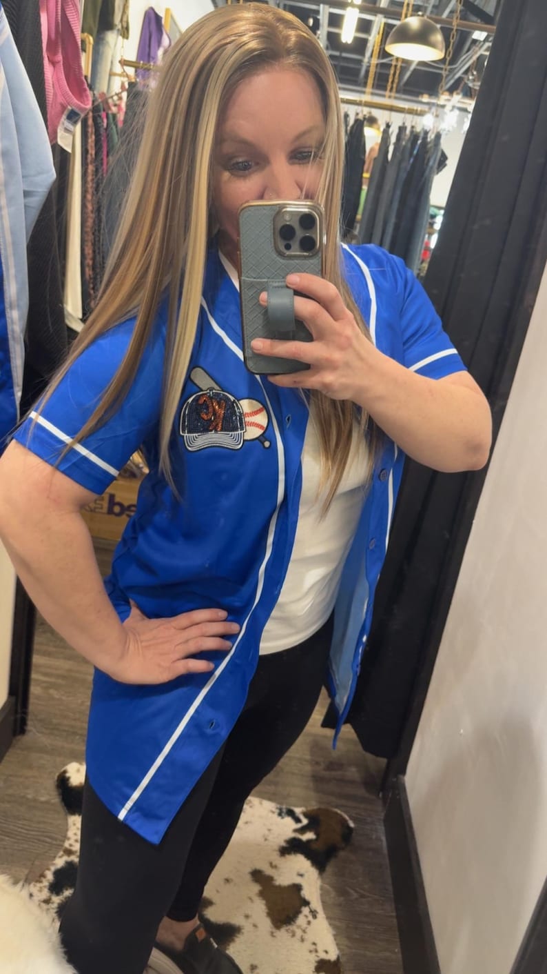Op de afbeelding: Een blauw baseballshirt met witte bies en een baseball-thema patch op de voorkant. Het shirt is knoopsluiting en heeft korte mouwen.