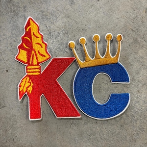 May include: A red and blue embroidered patch with the letters "KC" and a gold crown above the "C". The "K" has a red spearhead with a yellow feather design.