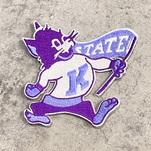 May include: Purple and white embroidered patch of a cartoon cat holding a flag that says "K-State".