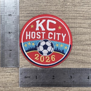 May include: A circular embroidered patch featuring a red background with the white text KC HOST CITY and the year 2026 in yellow. A soccer ball graphic sits above a blue landscape. A metal ruler shows the patch is approximately 3 inches in diameter.